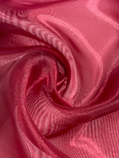 A close-up of smooth, flowing Organza - Jester Red fabric from Super Cheap Fabrics, 150cm wide, displays elegant swirling folds with a lustrous sheen and deep red hue, highlighting its delicate and luxurious quality.