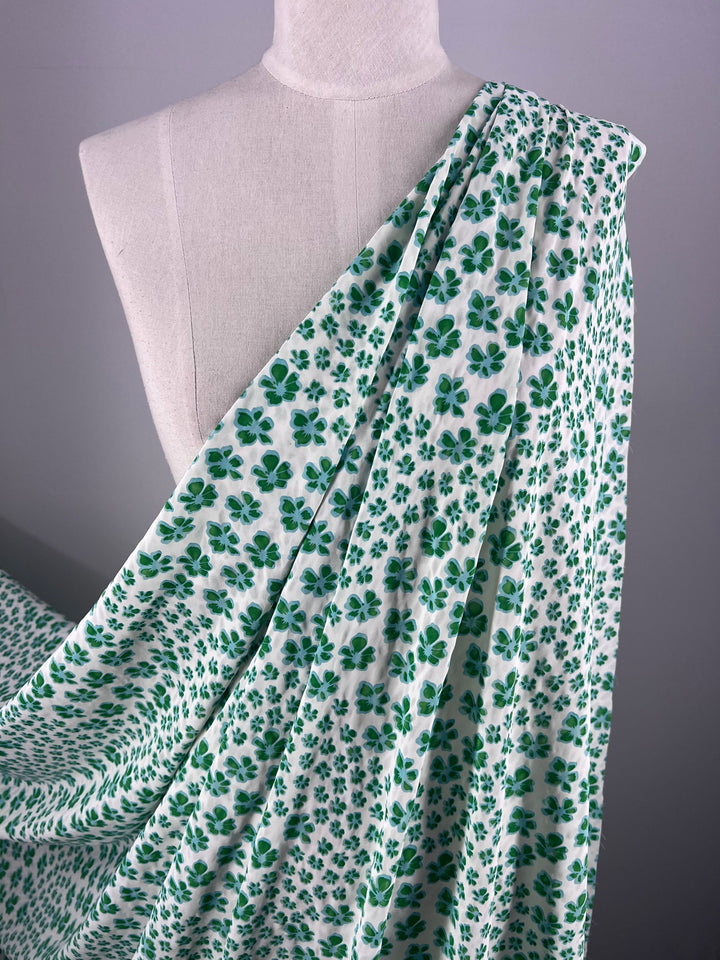 A mannequin showcases the Salt Shrunk Print - Smurf - 155cm by Super Cheap Fabrics, elegantly featuring a green clover pattern with vibrant colors cascading from one shoulder.