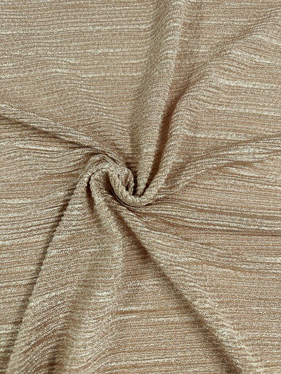 Close-up of Super Cheap Fabrics' Metallic Knit - Champagne, featuring a luxurious beige hue with a captivating metallic sheen. The fabric is displayed in a twisted pattern, showcasing its soft texture with slight crinkles and a blend of smooth and rippled surfaces. This medium-weight knit catches the light beautifully, enhancing its reflective and glittery quality. Width: 150cm.