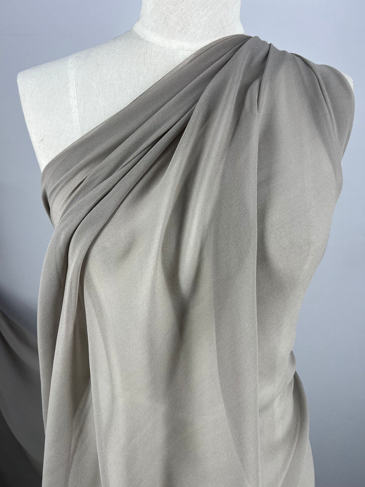 A mannequin is draped in a single piece of Silk Georgette - Earth from Super Cheap Fabrics, a light gray, sheer fabric that evokes the essence of spring and summer outfits. This 135cm wide material is arranged diagonally across the chest and shoulder, forming gentle folds and a flowing appearance. The plain background highlights the texture and color of the fabric beautifully.