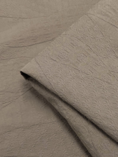 A close-up of Super Cheap Fabrics' Stretch Textured Bengaline Dune (150cm) reveals light taupe fabric with a subtle, abstract pattern. Overlapping layers highlight an intricate weave and soft, matte finish, enhanced with polyester for durability.