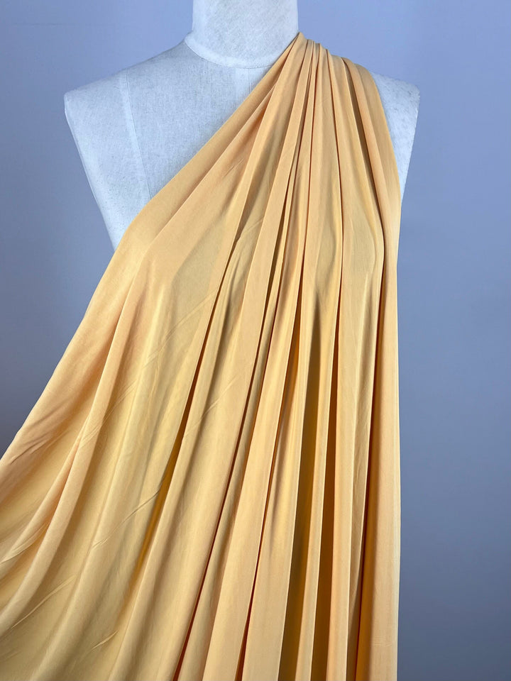 A mannequin draped with Super Cheap Fabrics' Summer Cotton Knit - Sunset Gold (160cm), covering one shoulder, creates a soft and elegant look. The pleated fabric highlights its smooth texture on a neutral background, enhancing the color.