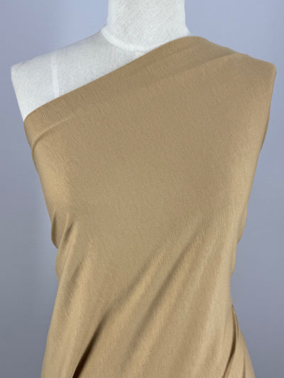 A mannequin showcases the Cotton Lycra - Doe by Super Cheap Fabrics, elegantly draped over one shoulder. The light brown fabric appears smooth with a subtle stretch. A plain light gray background enhances its soft allure.