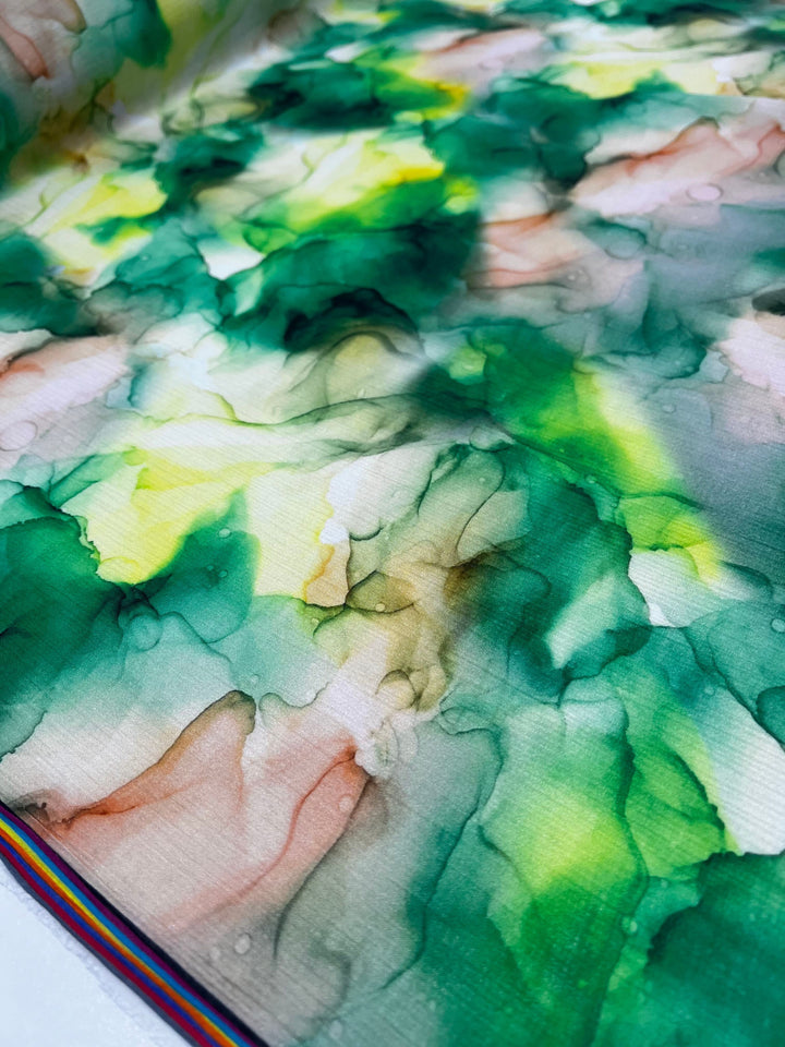 A close-up of the Designer Cotton - Fern - 150cm from Super Cheap Fabrics resembles a colorful abstract painting with fluid, marbled patterns in shades of green, yellow, and pink. The design includes a narrow stripe of rainbow colors along the lower edge, introducing a contrasting element to the organic forms.