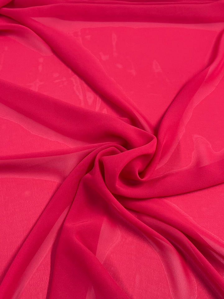 The Hi-Multi Chiffon - Hibiscus from Super Cheap Fabrics, with its vivid pink hue and sheer texture, gracefully drapes in soft folds to form a delicate spiral pattern.