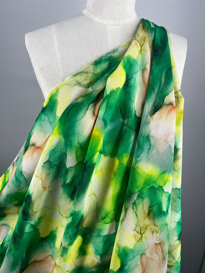 A mannequin dressed in luxurious sewing creations showcases a vibrant, lightweight fabric called Designer Cotton - Fern by Super Cheap Fabrics. This fabric features an abstract watercolor pattern in green, yellow, and white tones and flows gracefully over the shoulder, highlighting its colorful design.