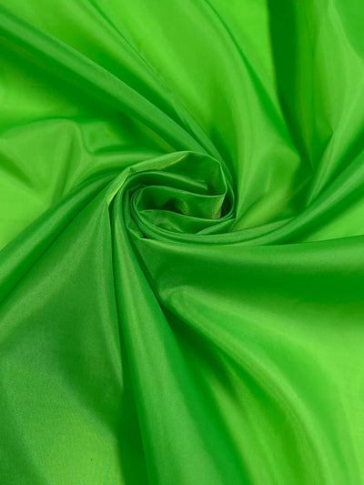 The Lining - Green Flash - 150cm by Super Cheap Fabrics features a close-up of vibrant green polyester fabric with a swirling pattern, creating movement and texture. The glossy sheen on its folds and creases highlights the smooth surface of this lightweight fabric.