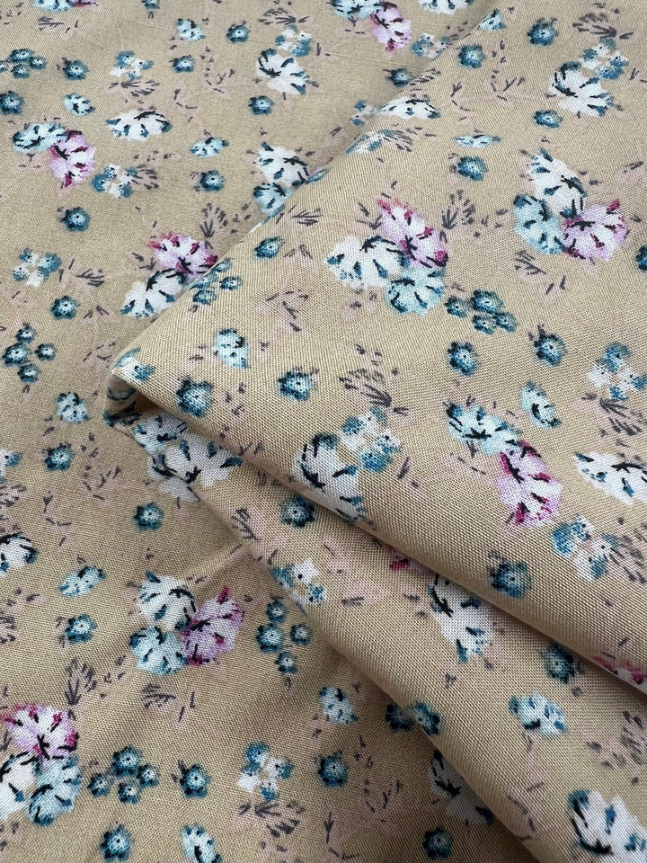 A close-up view of Super Cheap Fabrics' Printed Rayon - Pansy - 145cm, featuring a delicate floral pattern in shades of blue, purple, and white on a beige background. This versatile fabric is shown folded, revealing its vibrant prints on both visible layers.