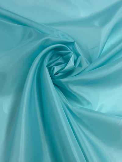 A close-up image of smooth, light blue satin fabric. The lightweight fabric is arranged in swirling folds, creating soft, gentle curves and a slightly glossy sheen that catches the light, highlighting its silky texture. This is the Lining - Angel Blue - 150cm by Super Cheap Fabrics.