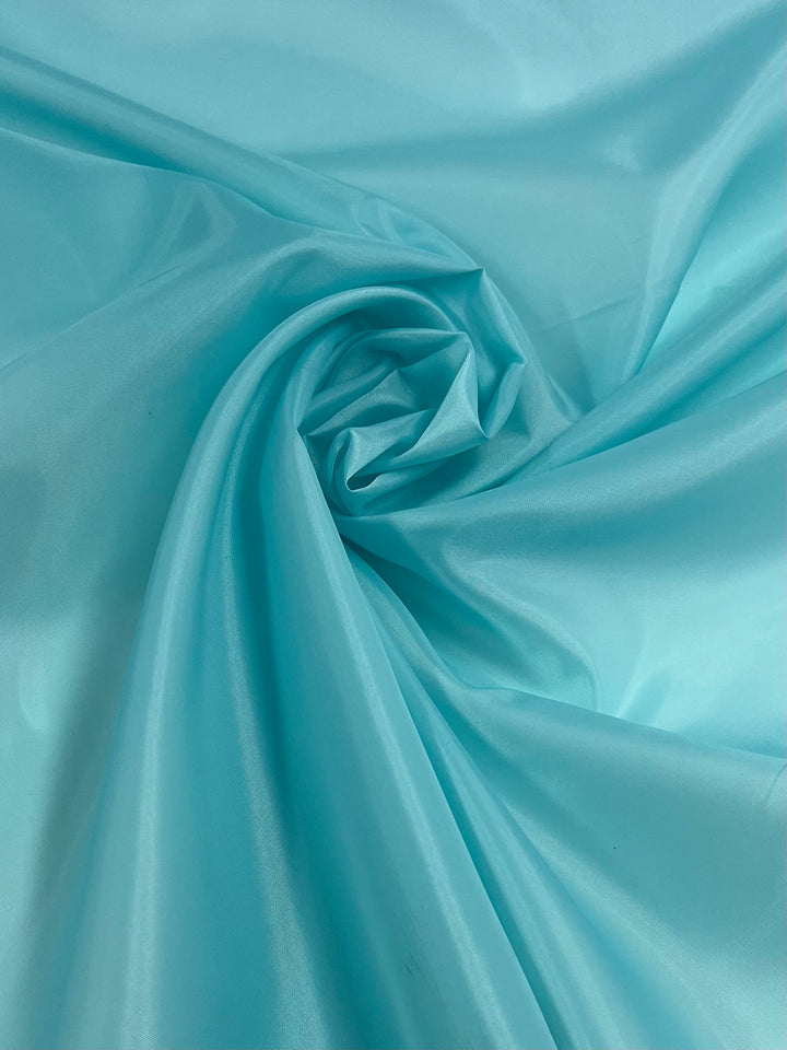 A close-up image of smooth, light blue satin fabric. The lightweight fabric is arranged in swirling folds, creating soft, gentle curves and a slightly glossy sheen that catches the light, highlighting its silky texture. This is the Lining - Angel Blue - 150cm by Super Cheap Fabrics.