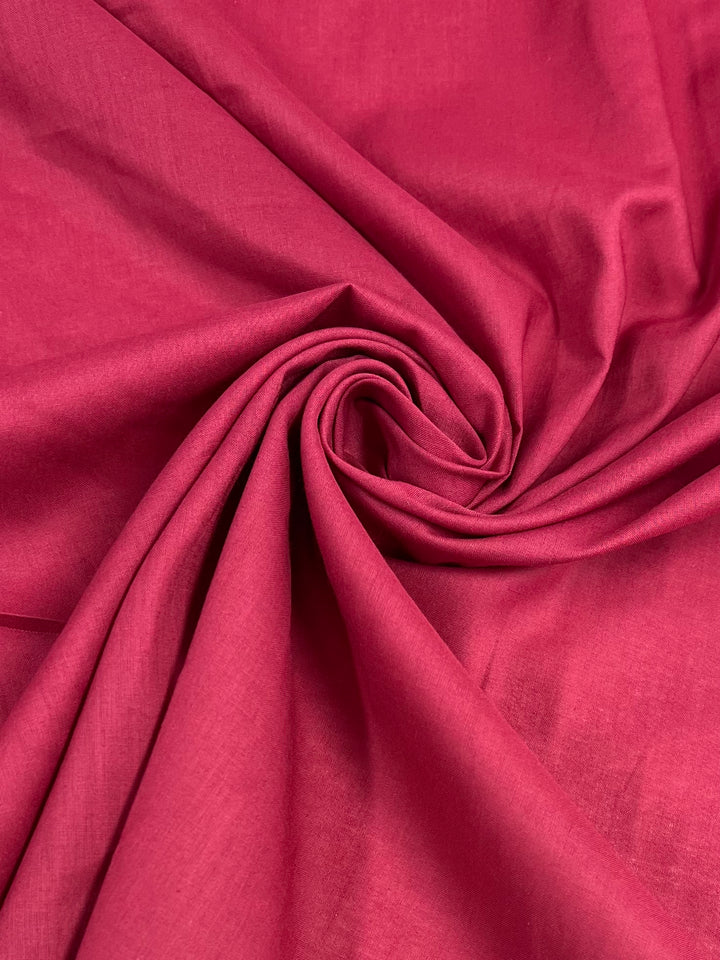 A close-up view of a piece of bright pink, extra light weight fabric arranged in a swirling, spiral pattern. The Cotton Voile - Jester Red - 140cm from Super Cheap Fabrics appears smooth and slightly glossy, with folds creating shadows that enhance the texture and depth.