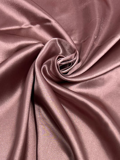 Close-up of a luxurious, smooth rose gold satin fabric perfect for dresses or home décor. The material is gathered in gentle folds and swirls, creating a glamorous, elegant texture with a soft, glossy sheen. This is the Satin - Ash Rose - 150cm from Super Cheap Fabrics.