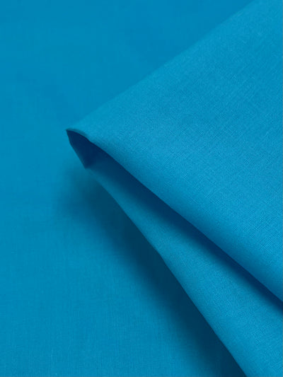 A close-up image of bright Cotton Voile - Blue Atoll - 140cm from Super Cheap Fabrics, neatly folded at a corner. The material appears smooth and lightweight, with a fine texture visible.