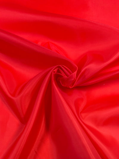 A close-up of Super Cheap Fabrics' Lining - Red - 150cm shows vibrant red fabric gathered in soft folds with a swirl pattern, silky sheen, and emerald green polyester lining that highlights its smooth, glossy surface.