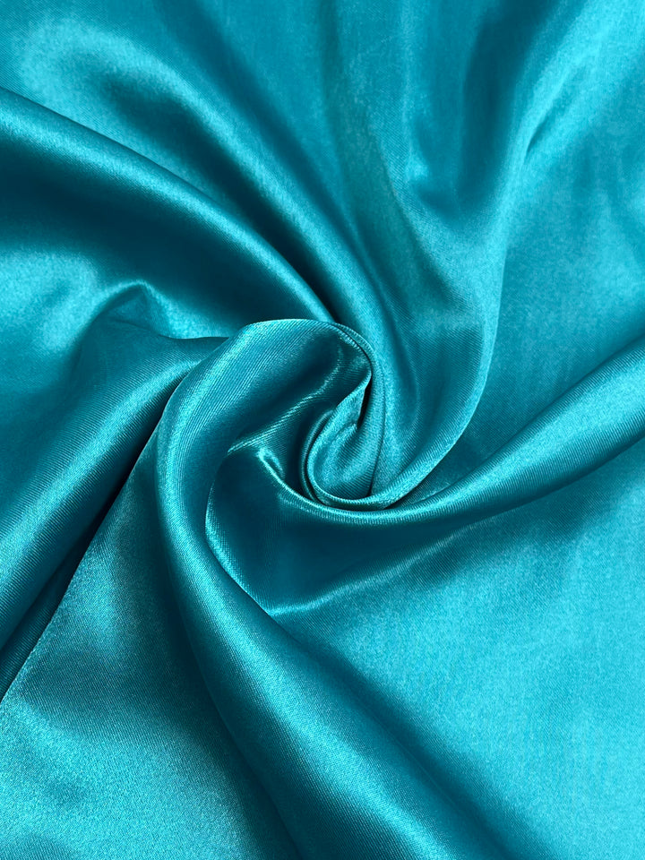 Close-up of a swirling, shiny teal satin fabric from Super Cheap Fabrics with a silky texture. The folds and creases create a dynamic flow, highlighting the lustrous surface and vibrant color of the Satin - Teal - 150cm polyester composition, making it an exquisite choice for elegant home décor.