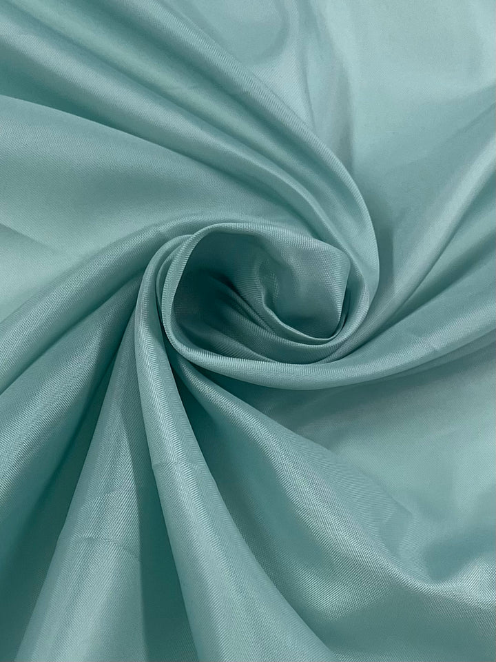 A close-up of a piece of Super Cheap Fabrics Lining - Egg Shell Blue - 120cm, twisted into a soft spiral pattern. The lightweight fabric appears smooth and slightly shiny, with gentle folds and creases creating a sense of texture and depth.