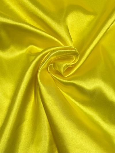 A close-up image of Super Cheap Fabrics Satin - Yellow - 150cm, perfect for dresses, skirts, tops, and accessories with a prominent swirl in the center forming soft, shiny folds. The fabric surface reflects light, enhancing its smooth texture and vibrant color.