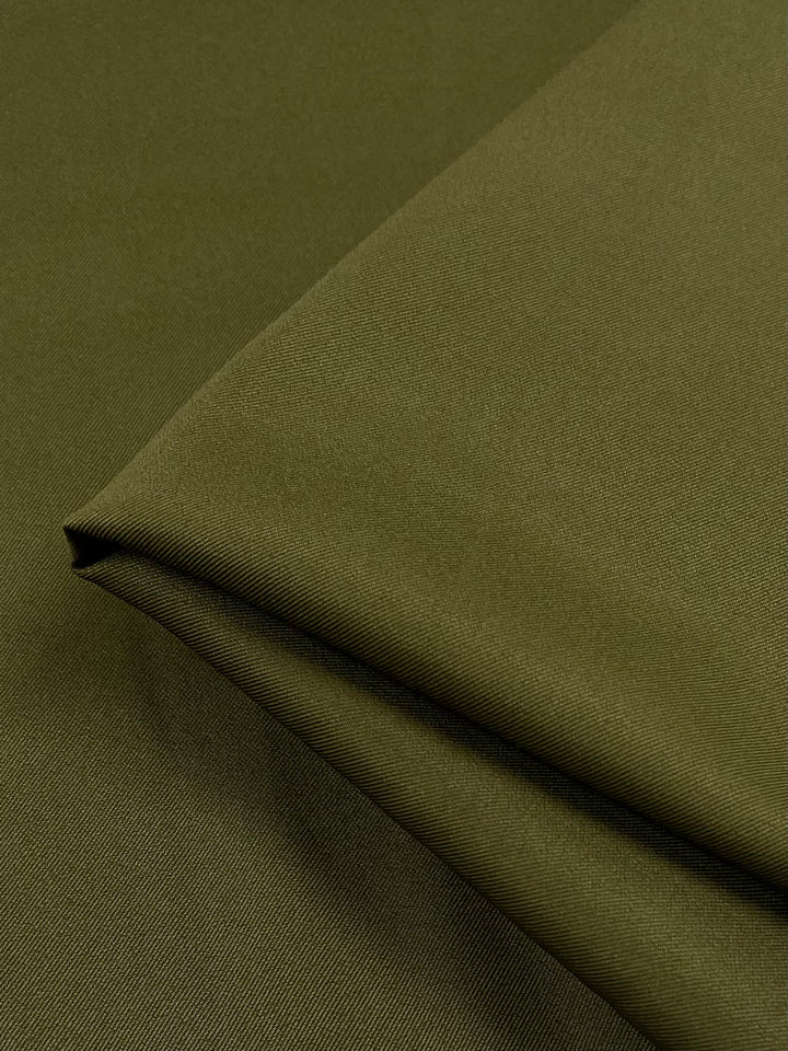 A close-up of a folded piece of Avocado Twill Suiting from Super Cheap Fabrics reveals its smooth, matte texture. Made from 100% polyester, the fabric is neatly folded to showcase clean lines and a subtle sheen. The uniform dark olive green color emphasizes the quality and versatility of this 155cm wide fabric.