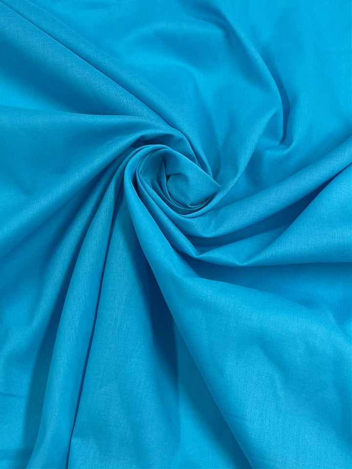 A close-up of Super Cheap Fabrics' Cotton Voile - Blue Atoll - 140cm swirled into a circular pattern at the center, creating folds and textures that radiate outward. The lightweight cotton material catches light, showcasing its soft and flowing nature.