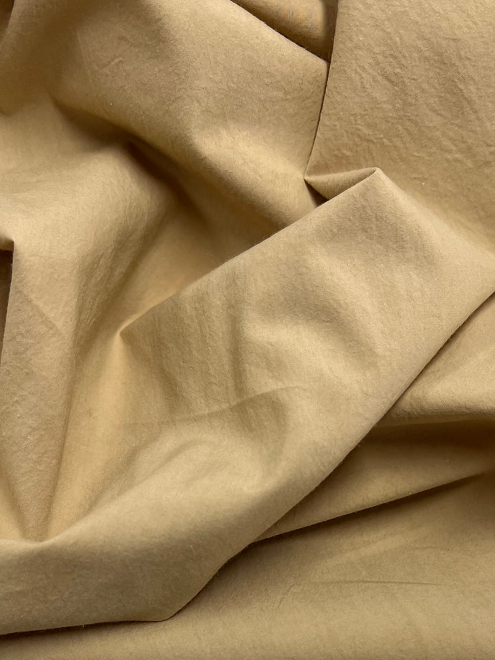 A close-up of sand-colored, crumpled fabric. The texture shows folds and creases, giving the 100% cotton material a slightly wrinkled appearance. The fabric appears soft and lightweight. This is Stretch Plain Cotton - Sand - 150cm from Super Cheap Fabrics.