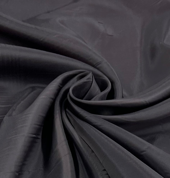 A close-up of Super Cheap Fabrics' Lining - Charcoal Art - 120cm reveals its smooth, shiny black polyester elegantly gathered into a swirling center, showcasing both texture and fluid drape.