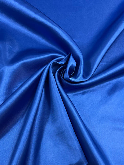 A close-up image of Super Cheap Fabrics' Satin - Royal - 150cm, showcasing its smooth, shiny texture. The fabric is arranged in soft folds and swirls, highlighting its lustrous surface and rich royal blue color—perfect for elegant dresses or sophisticated home décor accents.