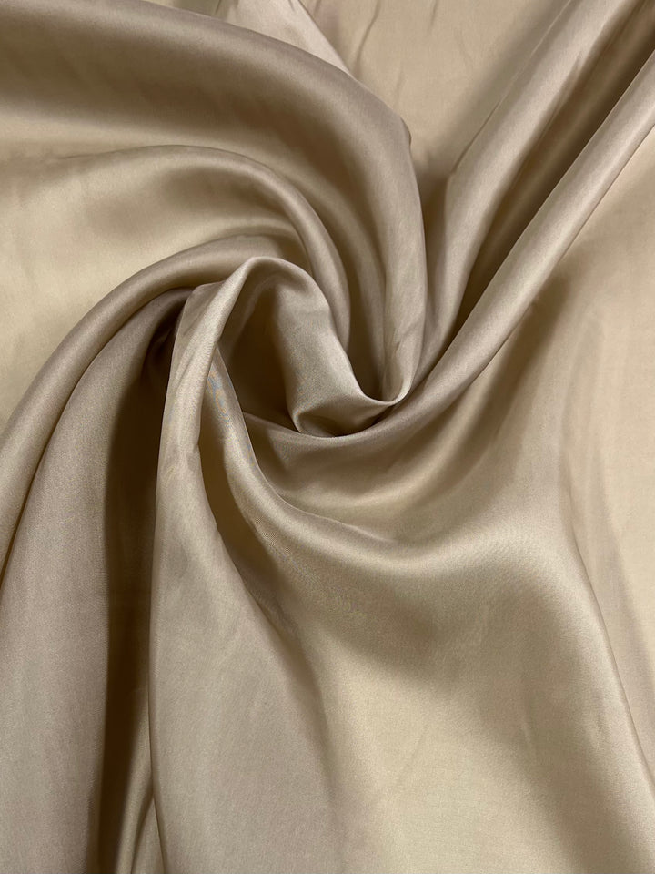 A close-up image of smooth, beige fabric artistically draped and twisted, creating elegant folds and flowing textures. Perfect for Jackets & Dresses, the material appears soft and slightly glossy, reflecting light subtly, adding depth and dimension to the image. The Lining - Ginger Root - 120cm from Super Cheap Fabrics truly enhances the elegance of this scene.