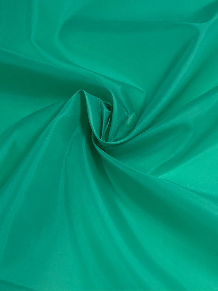 A close-up of a smooth, Aqua Green polyester lining with a swirl pattern in the center. The lightweight fabric appears glossy and flows gently from the center swirl, creating soft folds and creases. The vibrant shade of green is evenly distributed across the material. This is the Lining - Aqua Green - 150cm by Super Cheap Fabrics.