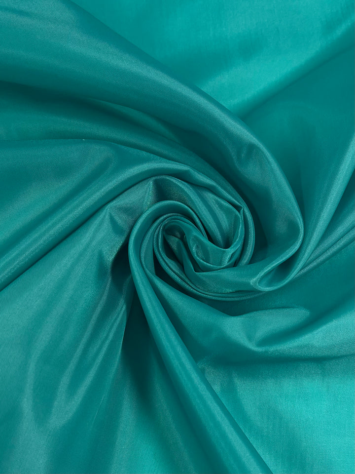 A close-up view of Super Cheap Fabrics' Lining - Teal - 150cm with a smooth, shiny texture, gathered and twisted at the center, creating soft folds and a swirling pattern. The fabric appears silky and vibrant, reflecting light to highlight its luxurious sheen—perfect for tailored silhouettes or adding elegant lining to any garment.