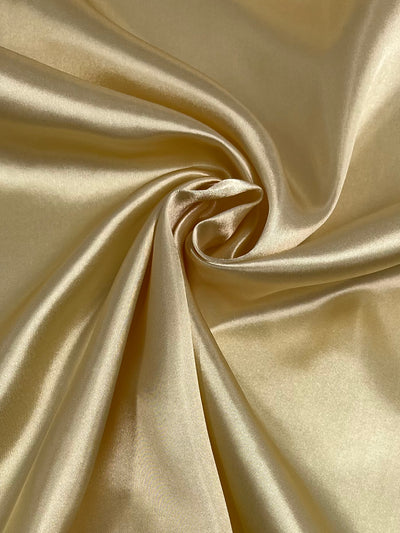 The Super Cheap Fabrics Satin - Italian Straw - 150cm features a smooth, luxurious golden fabric with swirling folds ideal for elegant dresses. Its shiny, reflective surface provides an exceptionally soft and exquisite appearance.