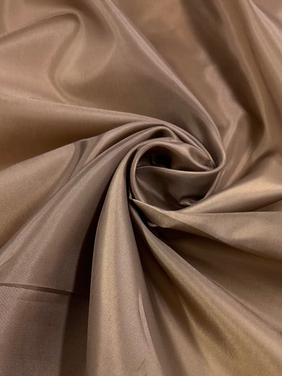 The Super Cheap Fabrics "Lining - Bronze Brown - 120cm" showcases a close-up of shiny polyester fabric, swirled into a spiral with smooth texture and radiating folds, emphasizing its lightweight silky appearance and subtle highlights.