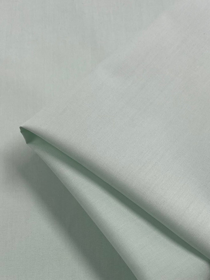 A smooth, folded piece of the Poplin - Ice Green fabric from Super Cheap Fabrics, made from light green polyester cotton with a subtle sheen, rests on a matching flat surface. This non-stretch fabric appears soft and lightweight, making it ideal for clothing or home textiles.