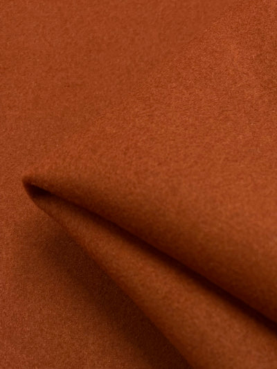 A close-up photo of a piece of rich, warm brown Wool Cashmere - Picante - 150cm fabric by Super Cheap Fabrics. The material is slightly folded, creating soft shadows and a sense of texture. The weave appears to be smooth, with a subtle sheen highlighting the luxurious wool.