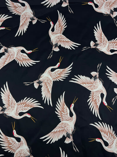 The Cotton Sateen - Fly High - 130cm by Super Cheap Fabrics features an elegant and dynamic design of white cranes with red crowns and yellow beaks, depicted in various flying poses with outstretched wings and long necks against a dark background. This medium-weight fabric is perfect for clothing projects.