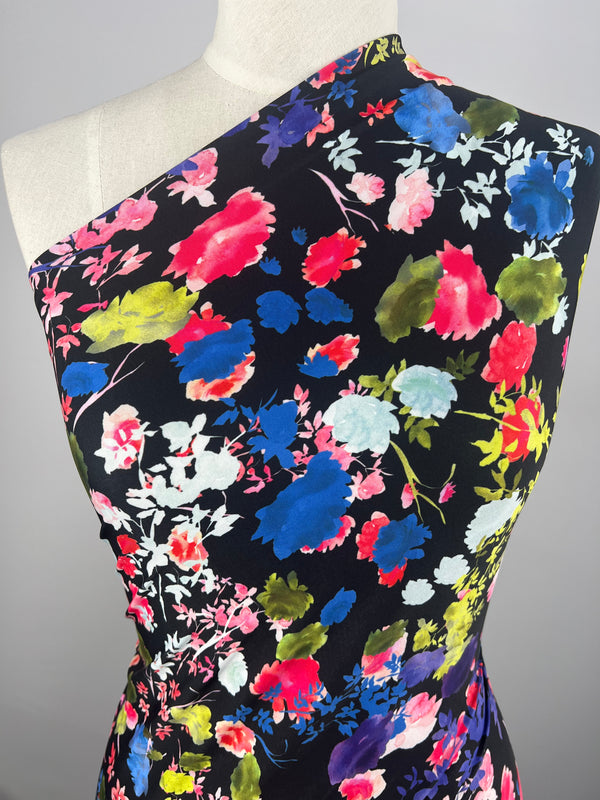 A mannequin is dressed in the Printed Lycra - Water Garden from Super Cheap Fabrics, showcasing a one-shoulder design with vibrant multicolored floral patterns on a black background. This medium-weight polyester fabric features flowers in shades of pink, blue, green, yellow, and white for a bold and lively multi-use garment.