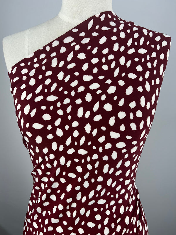 A mannequin showcases a chic and modern design with a maroon dress featuring a white abstract dot pattern created from the "Printed Lycra - Leopard Maroon - 150cm" fabric by Super Cheap Fabrics. The spandex blend allows for an elegant drape, enhancing the one-shoulder style.