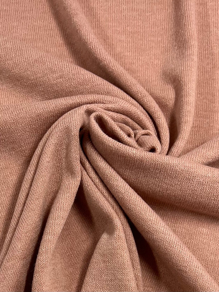 The **Textured Knit - Peach Melba - 145cm** fabric by **Super Cheap Fabrics** is shown in a close-up view, intricately gathered and twisted at the center to form a swirl pattern. The light brown or beige medium-weight material showcases a soft knit texture, perfect for crafting elegant dresses in the Peach Melba hue.