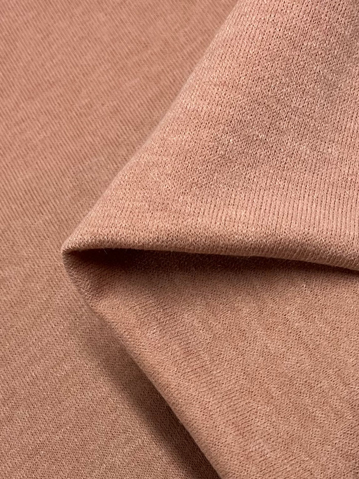 Close-up of a folded piece of Textured Knit - Peach Melba fabric from Super Cheap Fabrics. Its light brown, medium weight material showcases a soft texture and fine ribbing. Draped smoothly with a single visible fold creating depth and shadows, this 145cm knit fabric is perfect for crafting elegant dresses.