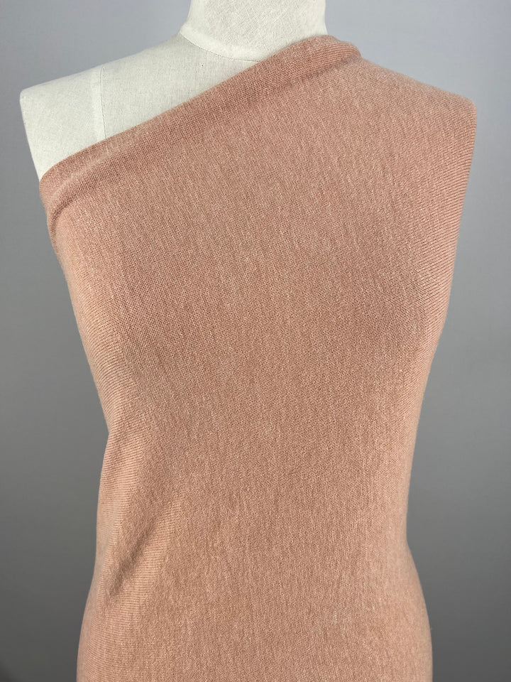 A mannequin is dressed in a one-shoulder garment made from Super Cheap Fabrics’ Textured Knit in Peach Melba. This simple and elegant top, crafted from medium-weight fabric, drapes smoothly over the mannequin, showcasing its soft texture. The plain neutral background emphasizes the garment's design.