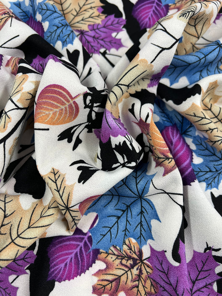 A close-up of a colorful polyester fabric with an autumn leaf pattern. The lightweight fabric features leaves in shades of orange, purple, blue, beige, and black against a white background. The Printed Crepe - Colourful Fall - 150cm from Super Cheap Fabrics is slightly wrinkled and folded in the middle, creating a textured appearance suitable for high fashion designs.