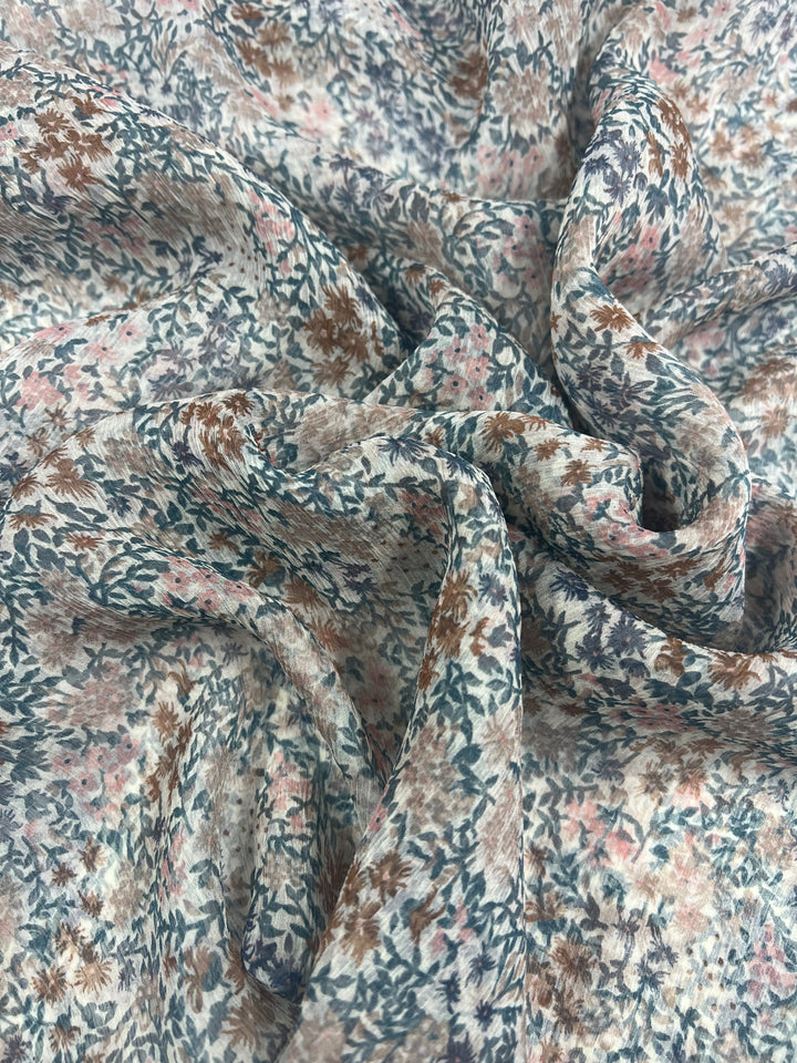 Close-up of a piece of luxury fabric with a detailed floral print featuring small flowers and leaves in muted tones of blue, brown, and pink on a light background. The Pure Printed Silk Chiffon - Soft Flowers by Super Cheap Fabrics is slightly crumpled, creating folds and shadows that add texture to the image.