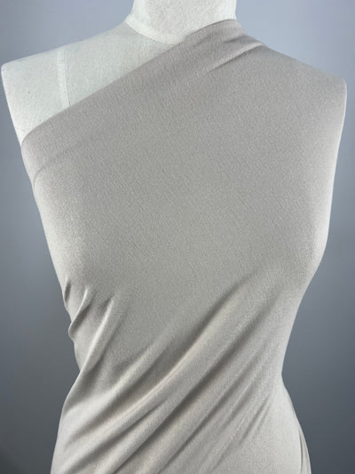 A white mannequin is draped in a beige, one-shoulder, 100% cotton fabric called Cotton Jersey - Oatmeal - 145cm from Super Cheap Fabrics. The material appears smooth and form-fitting, showcasing a minimalistic, elegant design. The background is a plain gray.