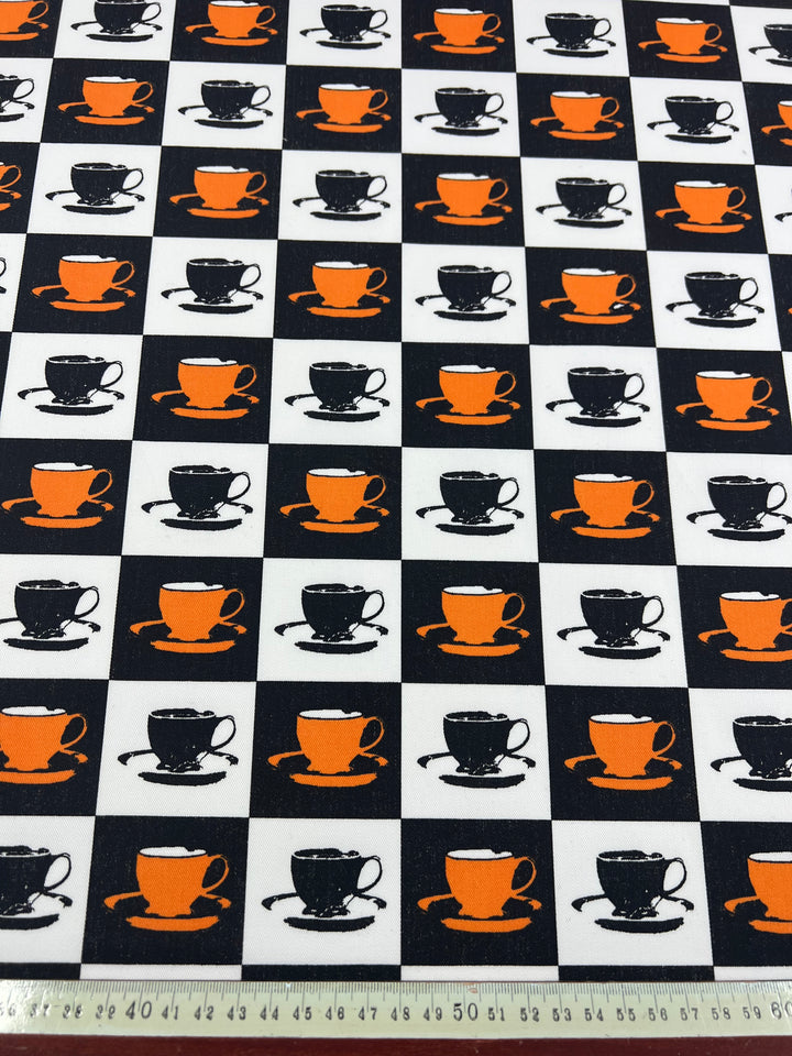 A 100% cotton drill fabric with a checkerboard pattern consisting of alternating black and white squares. Each square features a sketch-style illustration of a teacup; the cups in the black squares are orange, while the cups in the white squares are black. A measuring tape is at the bottom. This is **Super Cheap Fabrics' Cotton Drill - Orange MugKnight - 155cm**.