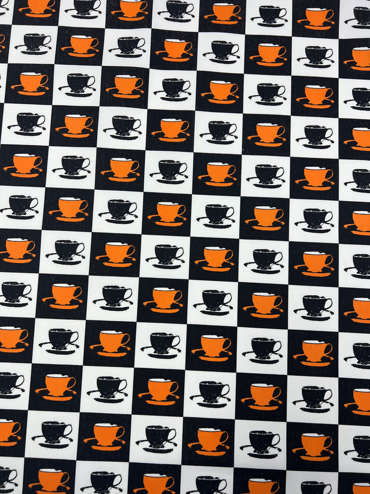 A repeating pattern featuring alternating black and orange coffee cups on white and black backgrounds, printed on 100% cotton drill fabric. Each square in the grid shows a coffee cup with a saucer, creating a checkerboard effect with durable diagonal weave for added style. Introducing Cotton Drill - Orange MugKnight - 155cm from Super Cheap Fabrics.