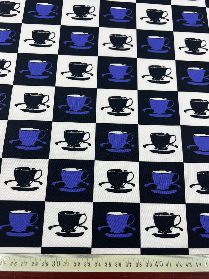 A lightweight cotton drill fabric featuring a checkerboard pattern of black and white squares. Each square contains an image of a tea cup and saucer in either blue (on black squares) or white (on white squares). The durable diagonal weave ensures longevity, with a ruler at the bottom displaying centimeters.

Introducing the **Cotton Drill - Purple MugKnight - 155cm** from **Super Cheap Fabrics**.