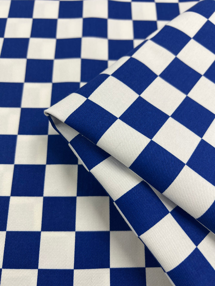 A close-up view of a piece of Cotton Drill - Blue Chess - 155cm by Super Cheap Fabrics with a blue and white checkerboard pattern. The fabric appears to be folded, emphasizing the alternating square design. The material has a slightly textured look, showcasing its durable diagonal weave.