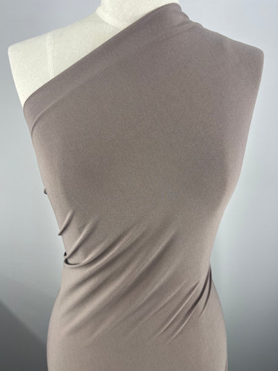 Close-up image of a dress form draped in a single-shouldered, taupe-colored ITY Knit - Mocha Meringue - 150cm fabric. The medium weight fabric by Super Cheap Fabrics is sleek, fitted, and has slight gathering, emphasizing its smooth texture and elegant flow. The background is plain and softly lit, highlighting the fabric's details.