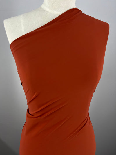 A mannequin draped in a sophisticated one-shoulder ensemble crafted from Super Cheap Fabrics' Nylon Lycra in Mango, measuring 145cm. This rust-colored attire showcases delicate draping and a four-way stretch for an elegant and fashionable look against a plain gray backdrop.