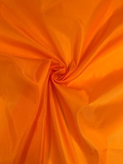 A vivid orange fabric, Lining -orange- 150cm from Super Cheap Fabrics, is gathered in the center, creating a spiral pattern with soft folds and creases radiating outward. The material appears smooth and slightly shiny, suggesting a lightweight, silky texture ideal as an accent for garments.