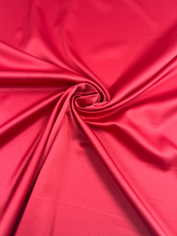 A close-up photo of the vibrant red fabric displays its smooth texture and subtle sheen. The lightweight stretch of Satin Deluxe - Red - 150cm from Super Cheap Fabrics is gathered in a swirl pattern at the center, highlighting its pliability and creating elegant folds and curves, perfect for evening wear.